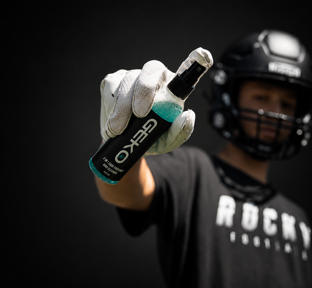 A person in a black football helmet and black shirt holds up a bottle of GEKO athletic shoe cleaner with white gloves, set against a dark background.