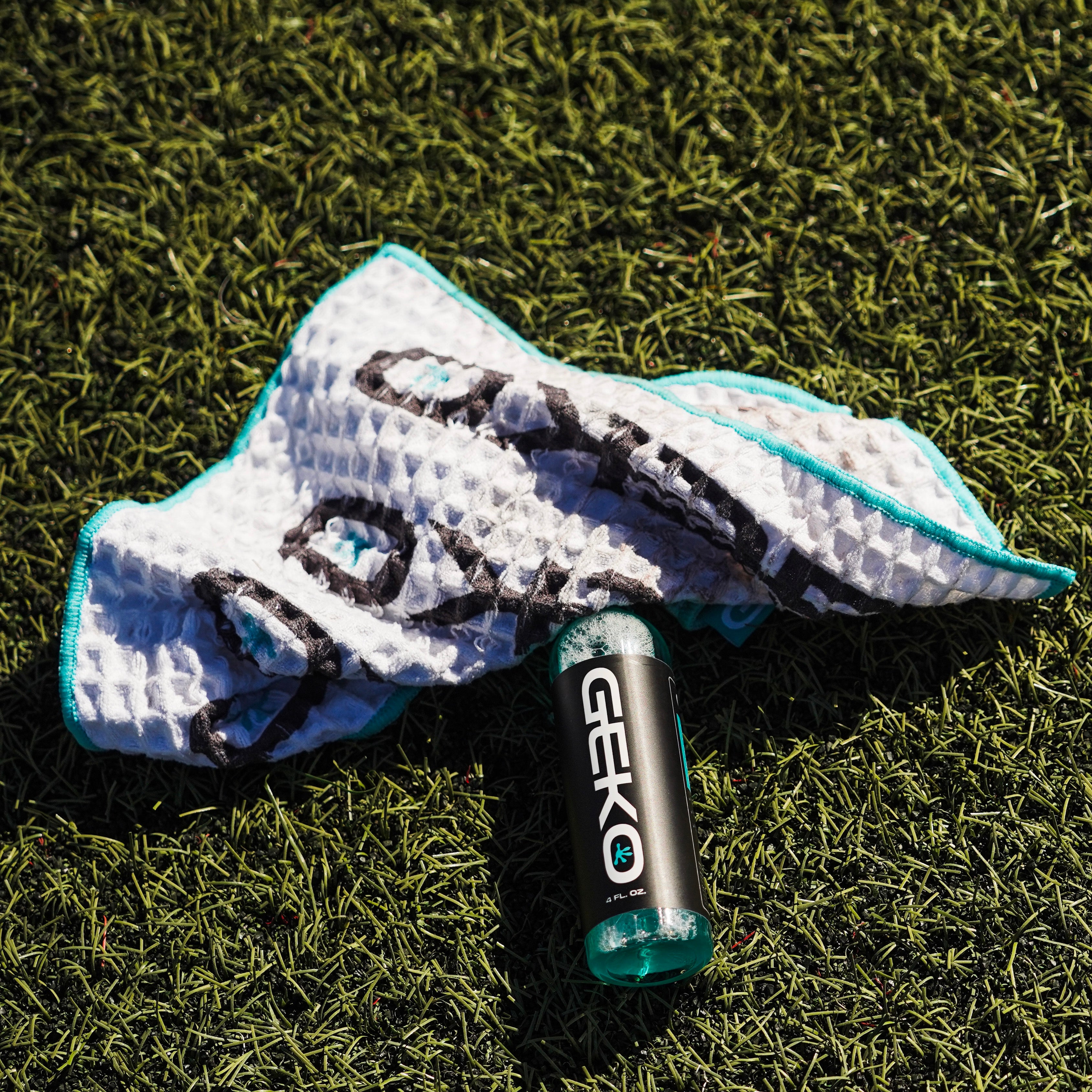 A GEKO sports cleaner bottle with clear teal liquid lies on a white textured towel with black and teal patterns, placed on green grass in bright sunlight.