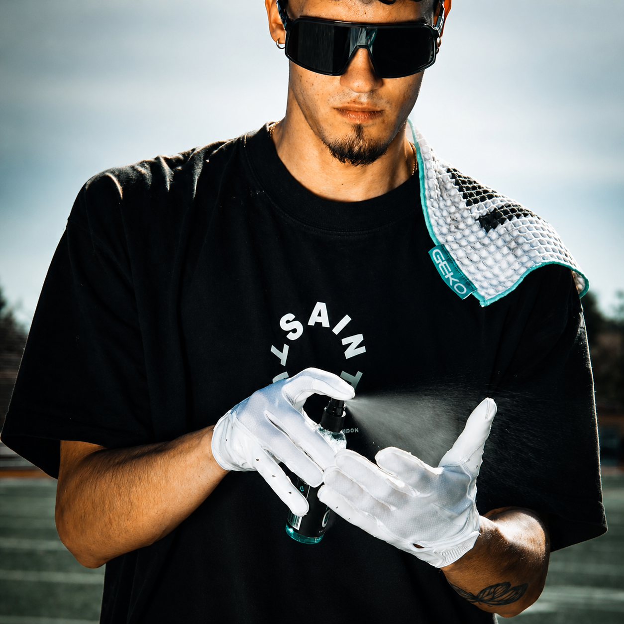 A man wearing a black t-shirt with a white circular graphic, white gloves, and black sunglasses sprays a bottle onto his hands. A teal and white GEKO cooling towel is draped over his shoulder, and the background shows a blurred outdoor setting.