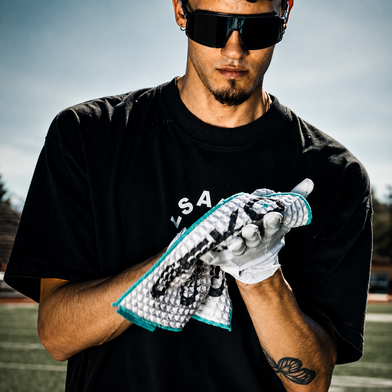 A man wearing a black t-shirt and black sunglasses on an outdoor sports field, holding a white athletic glove with a teal and black textured pattern.
